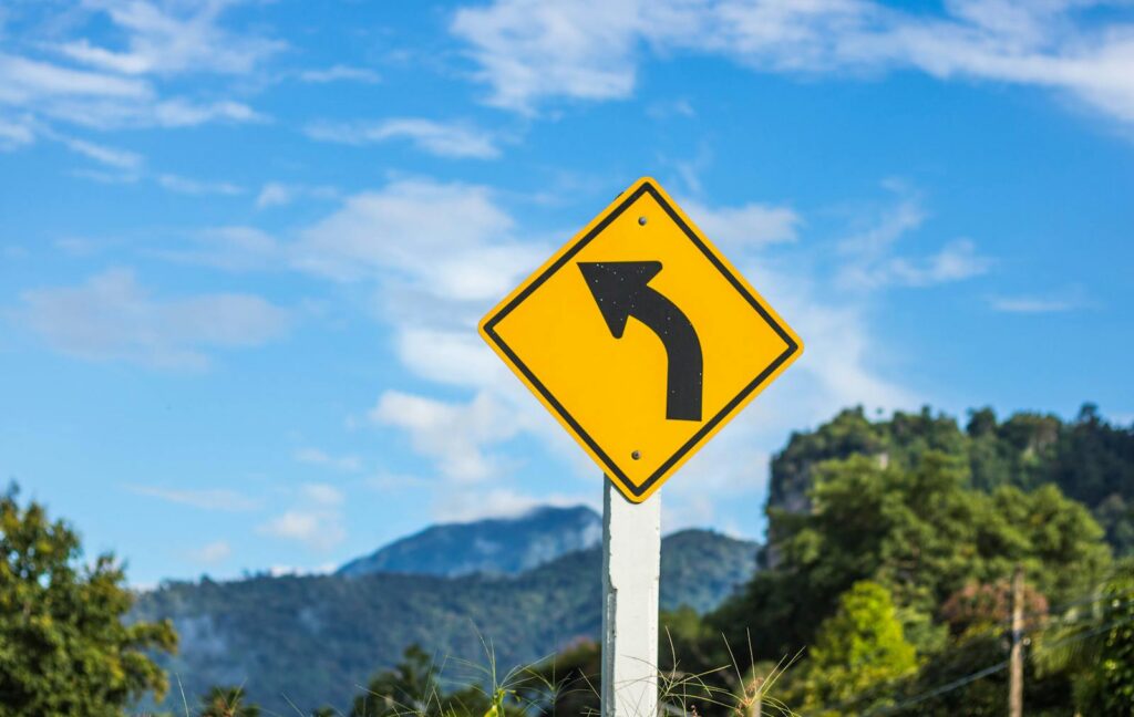 Yellow road sign with a left arrow against a scenic mountain backdrop on a clear day.