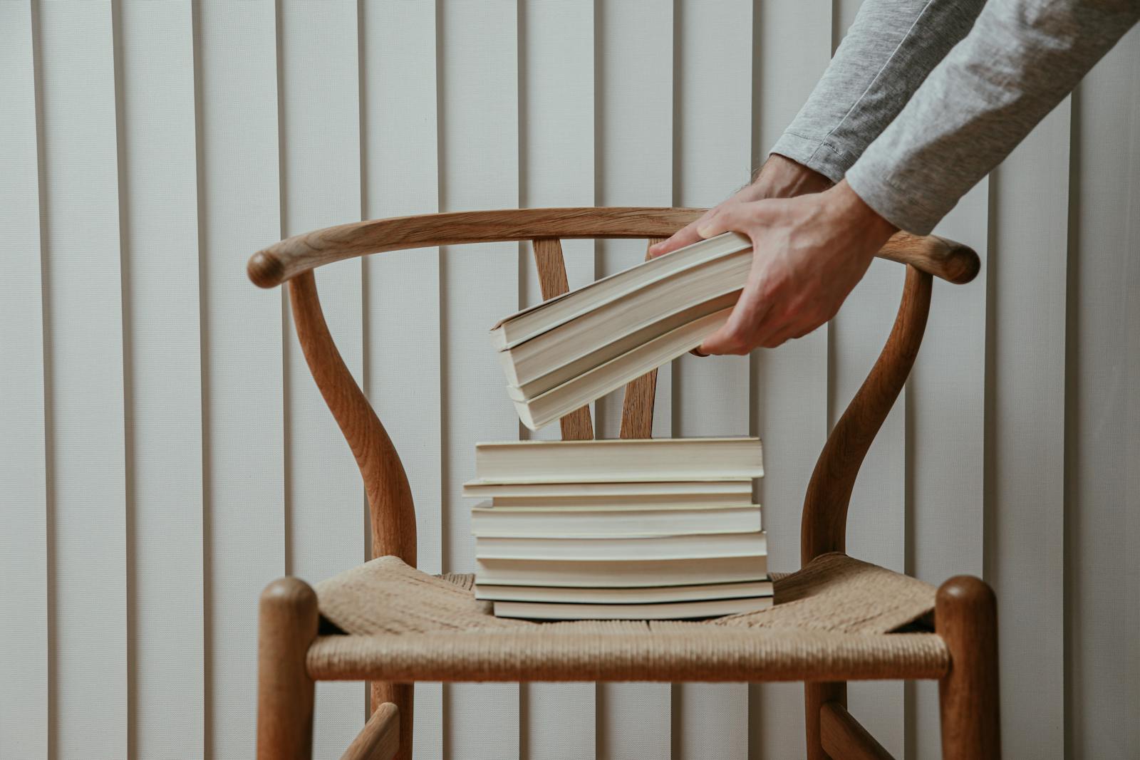 A person stacks books on a wooden chair against a white wall.